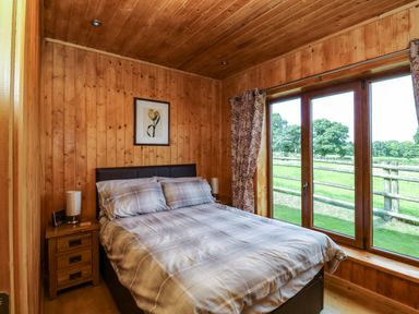 A bedroom with a double bed two bedside tables lamps and large window showing a garden view at Barn Shelley Lodge in Copplestone near Crediton