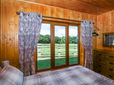 A bedroom with wooden walls a bed by a window with floral curtains and a dresser at Barn Shelley Lodge Copplestone near Crediton