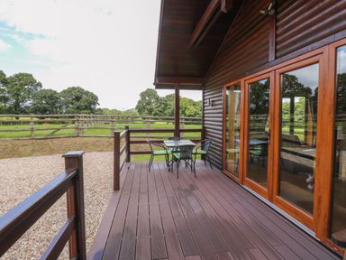 A wooden deck with a table and chairs overlooking a fenced grassy area at Barn Shelley Lodge in Copplestone near Crediton