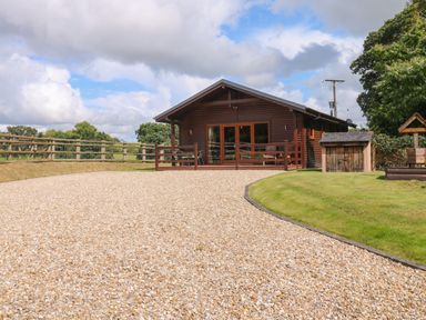 A gravel driveway leading to a wooden cabin with a fenced porch and outdoor furniture at Barn Shelley Lodge in Copplestone near Crediton
