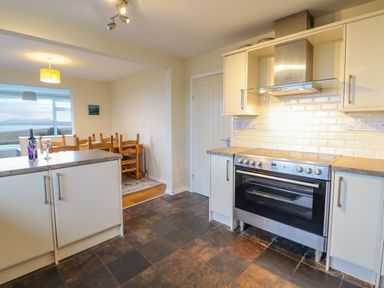 A kitchen with a stove and white cabinets and a dining area with wooden chairs and a window with a view at The Villa in Harlech