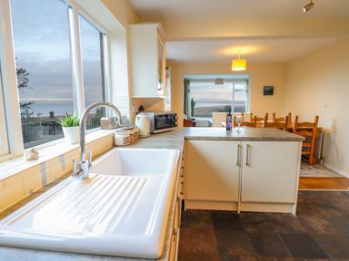 A kitchen with a white sink and faucet a countertop with a microwave kettle and jars and a dining area with wooden chairs and a window view at The Villa in Harlech