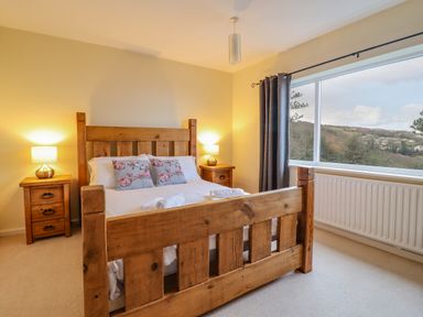 A bedroom with a wooden bed frame two bedside tables with lamps and a large window showing a view of hills and houses at The Villa in Harlech