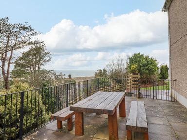 An outdoor patio with a wooden table and benches overlooking trees and the sea at The Villa in Harlech
