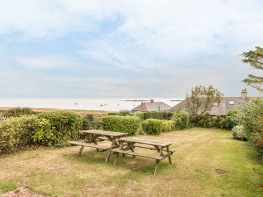 A garden with two wooden picnic tables and green bushes with houses and a sea view at 3B Coastguard Cottages in Low Newton-By-The-Sea