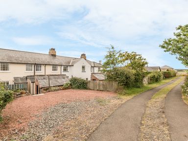 A row of white houses with chimneys next to a gravel driveway and a narrow paved road with greenery at 3B Coastguard Cottages in Low Newton-By-The-Sea