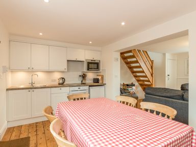 A kitchen and dining area with white cabinets a red and white checkered tablecloth on the dining table wooden chairs and a wooden staircase leading upstairs at 3B Coastguard Cottages in Low Newton-By-The-Sea