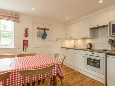 A kitchen with white cabinets a stove a sink a kettle wooden chairs and a table with a red and white checkered tablecloth at 3B Coastguard Cottages in Low Newton-By-The-Sea