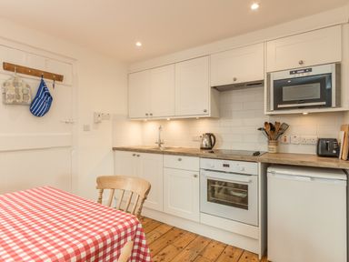 A kitchen with white cabinets a wooden chair a table with a red and white checkered tablecloth and various kitchen appliances at 3B Coastguard Cottages in Low Newton-By-The-Sea