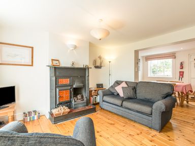 A living room with a dark grey sofa, armchair, fireplace, wooden floor and a dining area with a checkered tablecloth at 3B Coastguard Cottages in Low Newton-By-The-Sea
