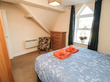 A bedroom with a bed covered by a blue blanket with towels on it a wooden chest of drawers a patterned armchair and a window with black curtains at Carisbrooke House Apartment 6 in Scarborough