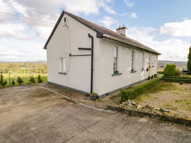 An exterior view of a white house with a sloped roof and multiple windows surrounded by a garden and concrete driveway at The Old School House in Gortermone near Carrigallen County Leitrim