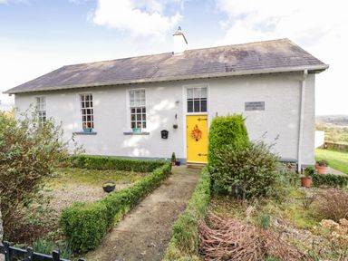 A single story white building with a yellow door and three windows surrounded by bushes and a garden path at The Old School House in Gortermone near Carrigallen County Leitrim