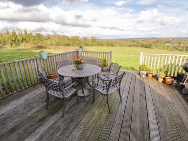 A wooden deck with a round table and six chairs surrounded by fence with plants on the railing and green fields beyond at The Old School House in Gortermone near Carrigallen County Leitrim