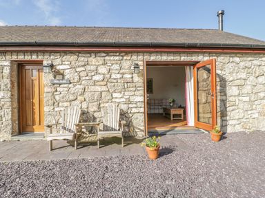 Exterior view of a stone building with wooden chairs outside and open door showing part of a living room at Hen Stabl Llangoed near Beaumaris