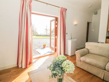 A living room with a sofa a coffee table with flowers wooden floors and open double doors with red and white striped curtains at Hen Stabl in Llangoed near Beaumaris
