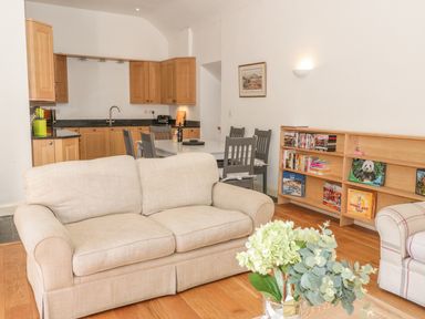A living room and kitchen area with beige sofa dining table with chairs and bookshelf at Hen Stabl in Llangoed near Beaumaris