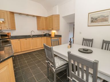 A kitchen with wooden cabinets a white dining table with gray chairs and a painting on the wall at Hen Stabl in Llangoed near Beaumaris