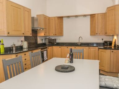 A kitchen with wooden cabinets black countertops a white table with chairs a bottle and food items on the table at Hen Stabl in Llangoed near Beaumaris