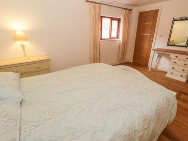 A bedroom with a bed a bedside table with a lamp a window with curtains a wooden door and a dressing table with a mirror at Hen Stabl in Llangoed near Beaumaris