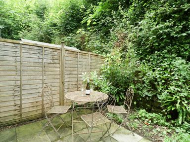 An outdoor patio area with a metal table and four chairs surrounded by a wooden fence and dense greenery at Wye Valley Cottage in Symonds Yat