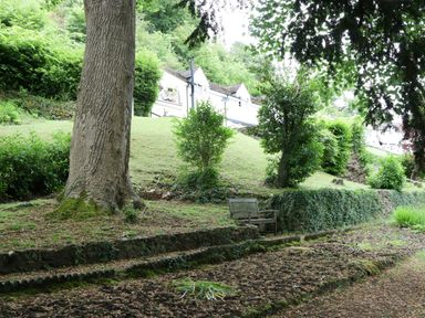 A garden with trees shrubs and a bench near a hillside with white houses at Wye Valley Cottage in Symonds Yat