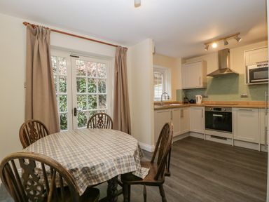A kitchen with a round table covered with a tablecloth and four chairs next to glass doors with curtains at Wye Valley Cottage in Symonds Yat