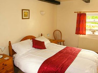 A bedroom with a bed with white linens and red accents a wooden chair and a window with a red curtain at Gorrig Bwthyn in Llandysul