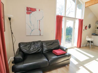 A black leather sofa with a red blanket folded on it next to tall windows with red curtains in a living area at Orchard Lodge in Warkworth