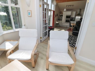 A sitting area with two white cushioned wooden chairs next to a window and a kitchen with dining table visible through open double glass doors at Orchard Lodge in Warkworth