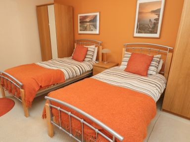 A bedroom with two single beds with orange blankets and pillows a wooden wardrobe a bedside table with a lamp and two framed pictures on an orange wall at Orchard Lodge in Warkworth