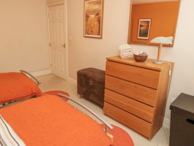 A bedroom with two beds covered with orange blankets a wooden chest of drawers with a lamp towels and decorative bowl a brown ottoman and a door on the wall at Orchard Lodge in Warkworth