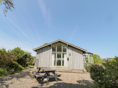 A wooden cabin with large windows and a picnic table outside surrounded by bushes at Orchard Lodge in Warkworth