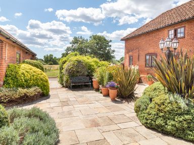 A patio with stone tiles surrounded by bushes and potted plants between two brick buildings at Horseshoe in Little Glemham near Wickham Market