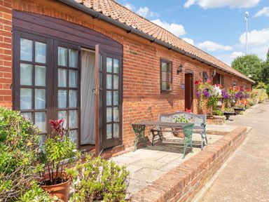A brick exterior of a building with French doors open to a patio with a metal bench and table and potted plants along a walkway at Horseshoe in Little Glemham near Wickham Market