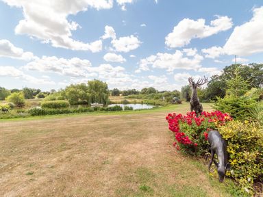 A garden with grass, red flowers, metal deer sculptures, a pond with a boat, and trees at Horseshoe in Little Glemham near Wickham Market