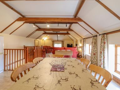 A dining area with wooden chairs and a table with a deer print tablecloth next to a sitting area with red sofas and exposed wooden beams at Hayloft Cottage in Little Glemham near Wickham Market