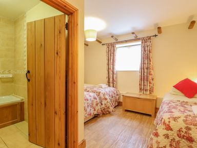 A bedroom with two single beds and a wooden chest near a window with curtains next to a wooden door leading to a tiled bathroom at Hayloft Cottage Little Glemham near Wickham Market