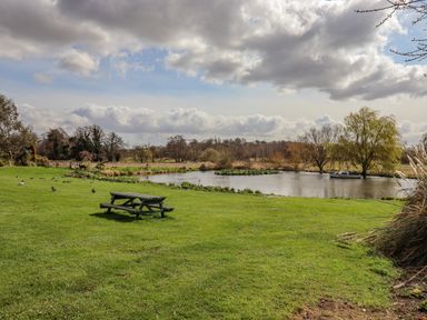 A grassy field with a wooden picnic bench near a pond with trees and a small boat at Hayloft Cottage in Little Glemham near Wickham Market
