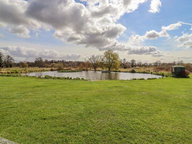 A pond surrounded by grass and trees with a small gazebo on the right at Hayloft Cottage in Little Glemham near Wickham Market