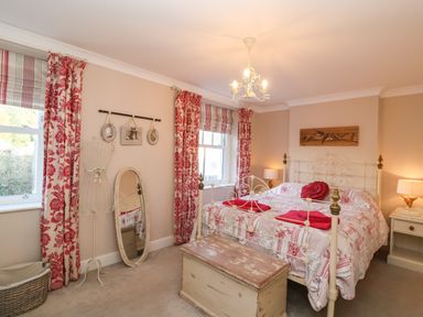 A bedroom with a bed with red and white bedding two windows with red floral curtains a wooden chest at the foot of the bed and a standing mirror at Enfield in Porlock