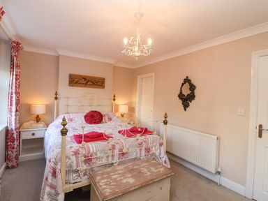 A bedroom with a bed with patterned bedding and red accents two bedside tables with lamps a wooden chest at the foot of the bed and a decorative mirror on the wall at Enfield in Porlock
