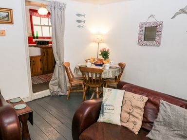 A dining area with a round table set with plates and glasses next to a brown leather sofa with cushions and a doorway to a kitchen at Inglenook Cottage in Whitby