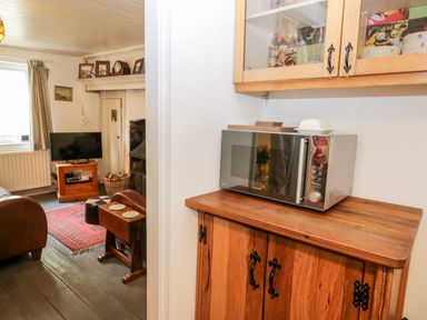 A kitchen area with a microwave on a wooden cabinet next to a living room with a television on a wooden stand and a fireplace at Inglenook Cottage in Whitby