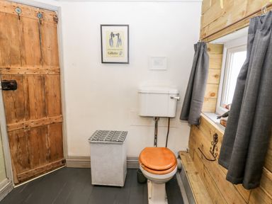 A bathroom with a wooden door, a toilet with a wooden seat, a white laundry basket, a window with gray curtains, and two framed pictures on the wall at Inglenook Cottage in Whitby
