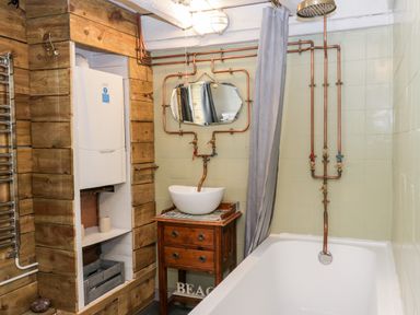 A bathroom with a bathtub shower combination a sink on a wooden dresser a wall mirror and exposed copper pipes at Inglenook Cottage Whitby