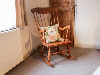 A wooden rocking chair with a floral cushion next to a window and wooden door at Inglenook Cottage Whitby