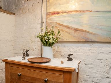 A wooden dresser with a white pitcher of flowers a round wooden plate and small decorative figurines against a white brick wall at Inglenook Cottage in Whitby