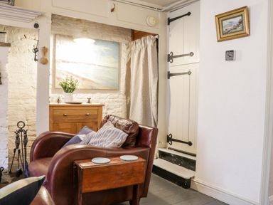 A living area with a brown leather armchair, wooden cabinet with a vase of flowers, a wall painting, and a white door at Inglenook Cottage in Whitby