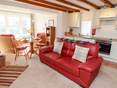 A living room with a red leather sofa two wooden chairs around a round wooden table and visible kitchen cabinets at The Bothy in Lowick near Holy Island